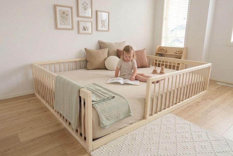 May include: A light wood-framed toddler bed with a mattress, surrounded by a railing. A child is sitting on the bed reading a book. The bed is decorated with pillows and a blanket. The room has a neutral color palette.