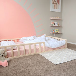 Puede incluir: Una cama para ni&ntilde;os peque&ntilde;os de madera clara con colch&oacute;n y ropa de cama rosas. Una alfombra blanca estampada est&aacute; en el suelo. Un oso de peluche y un juguete de peluche rosa est&aacute;n en el suelo cerca de la cama. La pared est&aacute; pintada de rosa con un dise&ntilde;o decorativo.