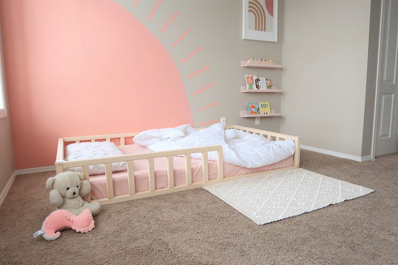Puede incluir: Una cama para ni&ntilde;os peque&ntilde;os de madera clara con colch&oacute;n y ropa de cama rosas. Una alfombra blanca estampada est&aacute; en el suelo. Un oso de peluche y un juguete de peluche rosa est&aacute;n en el suelo cerca de la cama. La pared est&aacute; pintada de rosa con un dise&ntilde;o decorativo.