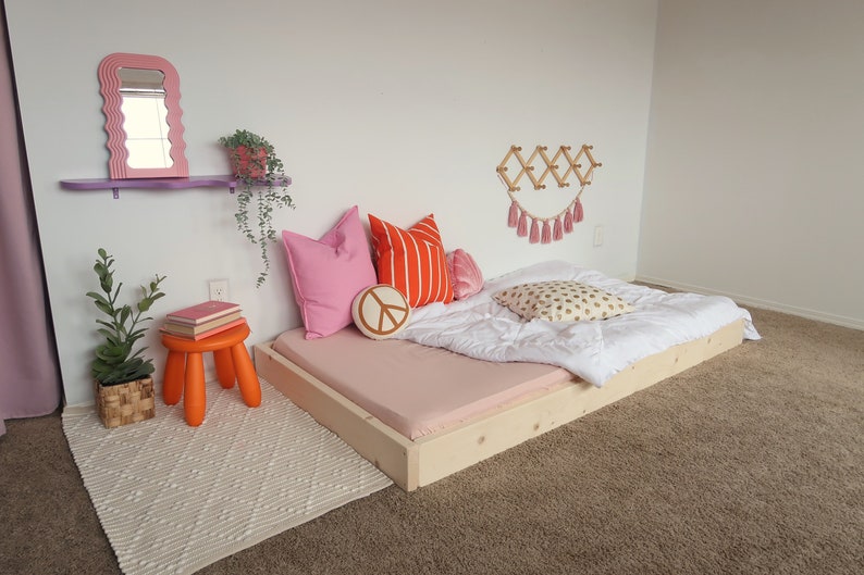 Puede incluir: Una cama baja con una s&aacute;bana bajera rosa claro, un edred&oacute;n blanco y cojines decorativos. Un peque&ntilde;o taburete naranja sostiene libros. Un espejo rosa con bordes ondulados y un perchero de pared de madera adornan la habitaci&oacute;n.