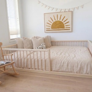 May include: A light wood rocking chair with a book sits next to a low-profile bed frame. The bed is made with a light-colored comforter and pillows. A framed sun art piece hangs above the bed, and a banner is strung across the wall.