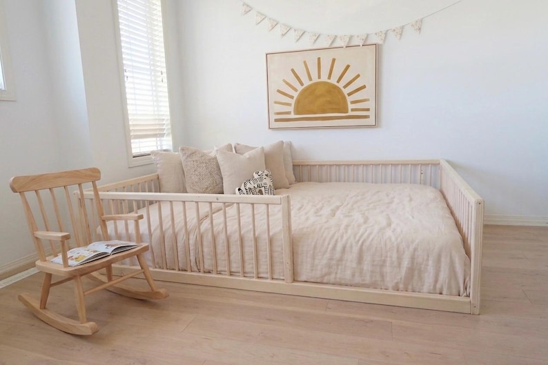May include: A light wood rocking chair with a book sits next to a low-profile bed frame. The bed is made with a light-colored comforter and pillows. A framed sun art piece hangs above the bed, and a banner is strung across the wall.