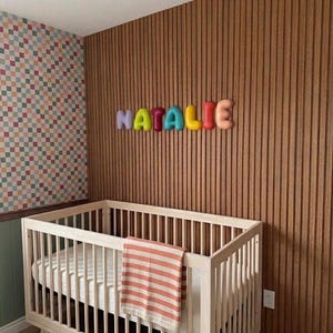 May include: A light-colored wooden crib with a striped orange and white blanket draped over the side. Above the crib, the name NATALIE is displayed in colorful, rounded, three-dimensional letters mounted on a vertical wood slat wall.