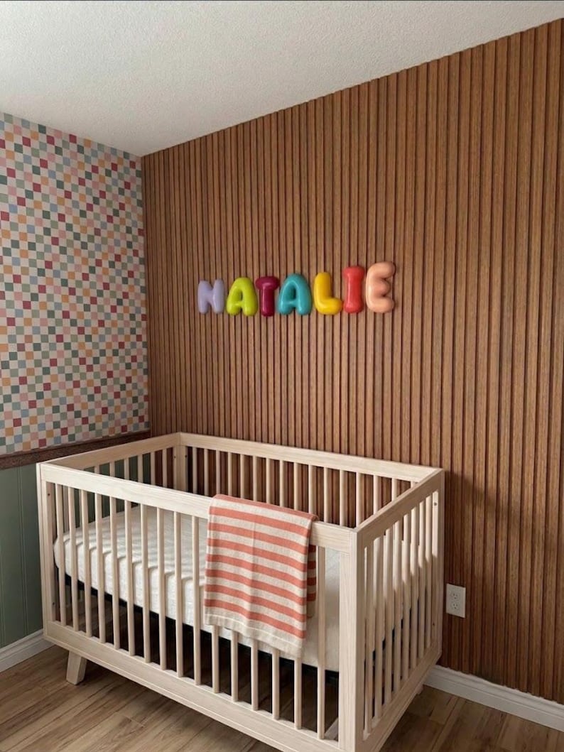 May include: A light-colored wooden crib with a striped orange and white blanket draped over the side. Above the crib, the name NATALIE is displayed in colorful, rounded, three-dimensional letters mounted on a vertical wood slat wall.