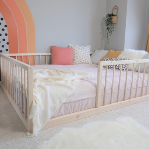 May include: A light-colored wooden bed frame with a low profile, surrounded by a railing. The bed is made up with a pink sheet, a cream-colored blanket, and several decorative pillows. A rainbow mural is painted on the wall.