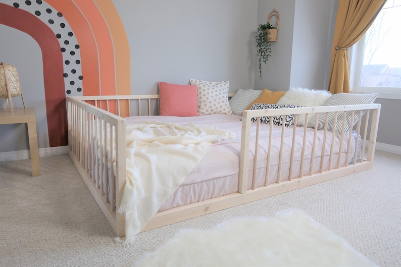 May include: A light-colored wooden bed frame with a low profile, surrounded by a railing. The bed is made up with a pink sheet, a cream-colored blanket, and several decorative pillows. A rainbow mural is painted on the wall.