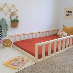 Puede incluir: Cama infantil de madera natural con una funda de colch&oacute;n color terracota. La habitaci&oacute;n cuenta con cojines decorativos, una almohada con una carita sonriente y un letrero que dice "YOU ARE MY SUNSHINE". La cama mide unos 160 cm de largo.