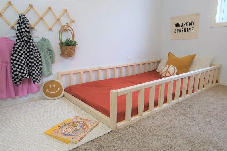 Puede incluir: Cama infantil de madera natural con una funda de colch&oacute;n color terracota. La habitaci&oacute;n cuenta con cojines decorativos, una almohada con una carita sonriente y un letrero que dice "YOU ARE MY SUNSHINE". La cama mide unos 160 cm de largo.