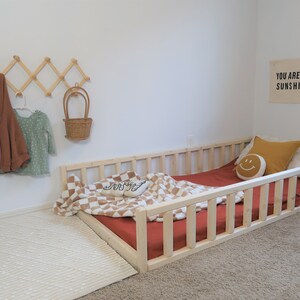 Puede incluir: Una cama baja para ni&ntilde;os peque&ntilde;os de madera clara con un colch&oacute;n rojo y una manta a cuadros. Un perchero de pared de madera sostiene ropa de ni&ntilde;os y una cesta de mimbre. Un letrero que dice "YOU ARE MY SUNSHINE" cuelga de la pared.