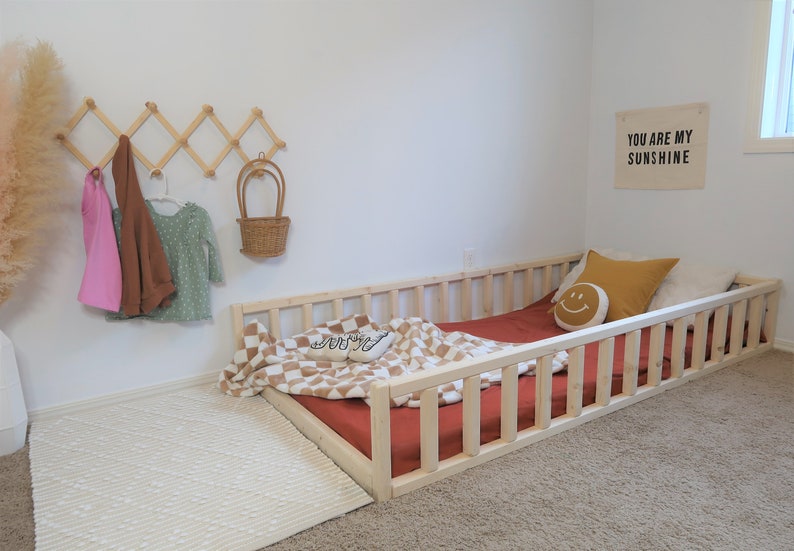 Puede incluir: Una cama baja para ni&ntilde;os peque&ntilde;os de madera clara con un colch&oacute;n rojo y una manta a cuadros. Un perchero de pared de madera sostiene ropa de ni&ntilde;os y una cesta de mimbre. Un letrero que dice "YOU ARE MY SUNSHINE" cuelga de la pared.