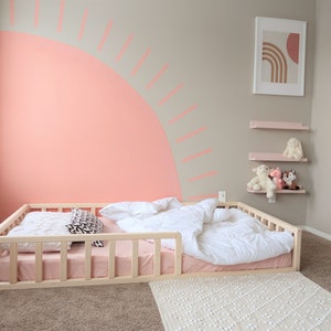 Puede incluir: Dormitorio infantil con una cama baja de madera, un colch&oacute;n rosa y ropa de cama blanca. La pared est&aacute; pintada con un gran dise&ntilde;o de sol color coral. Estanter&iacute;as rosas sostienen peluches y una impresi&oacute;n art&iacute;stica enmarcada cuelga.