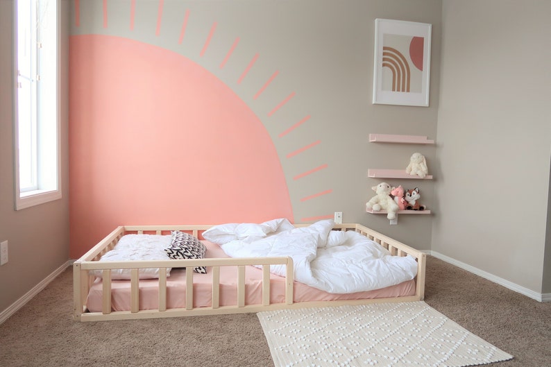 Puede incluir: Dormitorio infantil con una cama baja de madera, un colch&oacute;n rosa y ropa de cama blanca. La pared est&aacute; pintada con un gran dise&ntilde;o de sol color coral. Estanter&iacute;as rosas sostienen peluches y una impresi&oacute;n art&iacute;stica enmarcada cuelga.