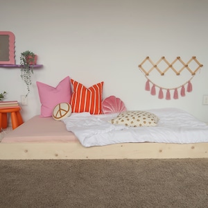 Puede incluir: Una cama baja de madera con una s&aacute;bana bajera rosa, un edred&oacute;n blanco y cojines decorativos. Los cojines incluyen rosa liso, rayas naranjas, un signo de la paz y una concha. Tambi&eacute;n se ven un taburete naranja peque&ntilde;o y un espejo rosa.