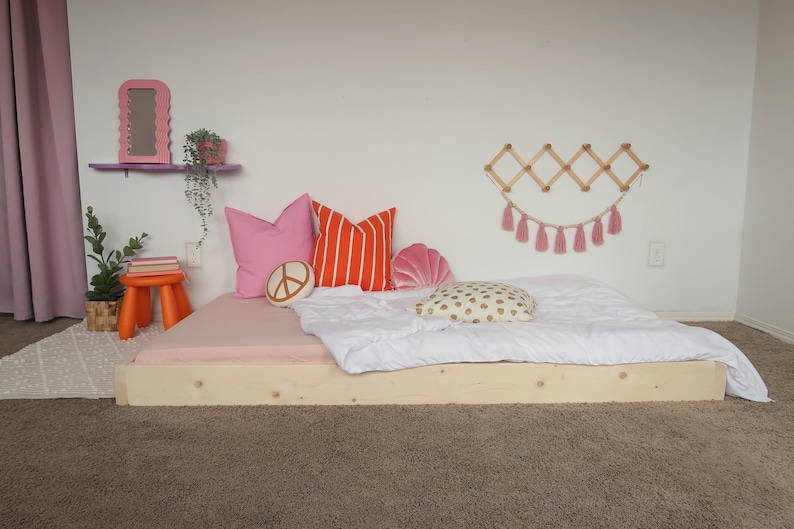 Puede incluir: Una cama baja de madera con una s&aacute;bana bajera rosa, un edred&oacute;n blanco y cojines decorativos. Los cojines incluyen rosa liso, rayas naranjas, un signo de la paz y una concha. Tambi&eacute;n se ven un taburete naranja peque&ntilde;o y un espejo rosa.