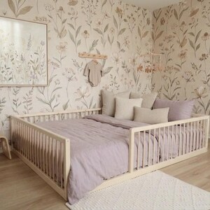 May include: A light wood framed bed with a lavender comforter and pillows. The room has floral wallpaper and a small side table with a basket. A framed floral print hangs on the wall.