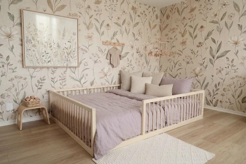 May include: A light wood framed bed with a lavender comforter and pillows. The room has floral wallpaper and a small side table with a basket. A framed floral print hangs on the wall.