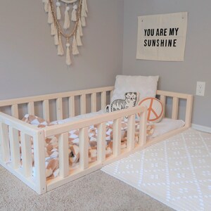 Puede incluir: Una cama para ni&ntilde;os peque&ntilde;os de madera natural con un perfil bajo y laterales de listones. La cama est&aacute; amueblada con una manta estampada y almohadas. Es visible un tapiz de pared con el texto "YOU ARE MY SUNSHINE".