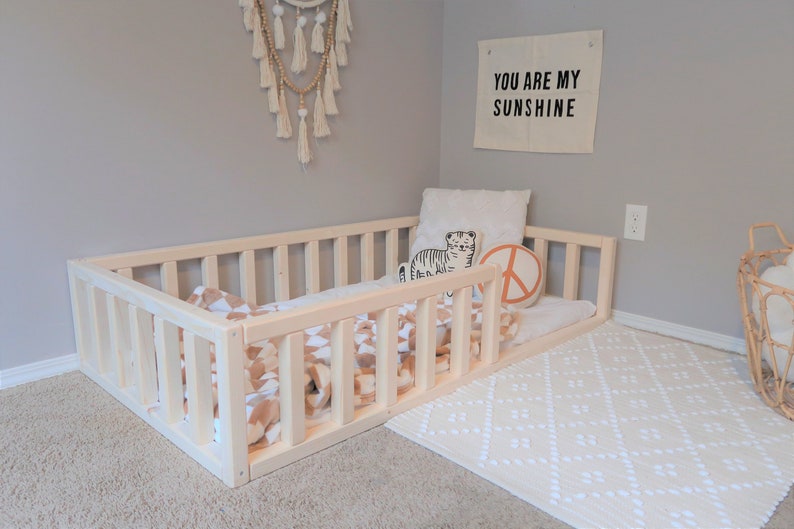 Puede incluir: Una cama para ni&ntilde;os peque&ntilde;os de madera natural con un perfil bajo y laterales de listones. La cama est&aacute; amueblada con una manta estampada y almohadas. Es visible un tapiz de pared con el texto "YOU ARE MY SUNSHINE".