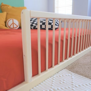 May include: A light-colored wooden crib with a red sheet. The crib has vertical bars on the side. Yellow pillows and a peace sign pillow are on the bed. A black and white checkered pillow is also on the bed.