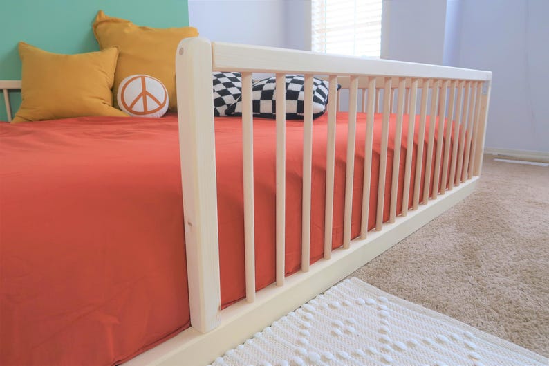 May include: A light-colored wooden crib with a red sheet. The crib has vertical bars on the side. Yellow pillows and a peace sign pillow are on the bed. A black and white checkered pillow is also on the bed.