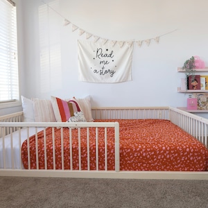 May include: A light wood-framed bed with a low profile, featuring a rust-colored blanket with a pink floral pattern. The bed is adorned with pillows and a decorative banner that reads "Read me a story."