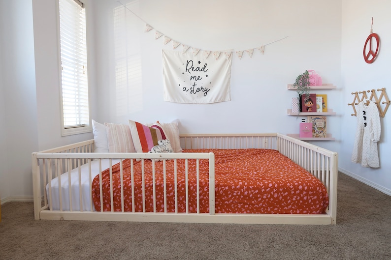 May include: A light wood-framed bed with a low profile, featuring a rust-colored blanket with a pink floral pattern. The bed is adorned with pillows and a decorative banner that reads "Read me a story."