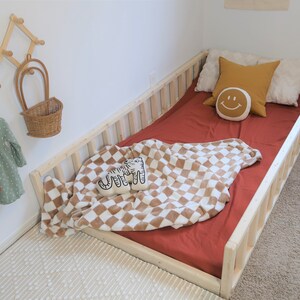Puede incluir: Una cama infantil con estructura de madera natural y una s&aacute;bana bajera de color &oacute;xido. La cama est&aacute; adornada con almohadas, una manta a cuadros y un coj&iacute;n decorativo con una cara sonriente. Un perchero acorde&oacute;n de madera y una peque&ntilde;a cesta cuelgan de la pared.
