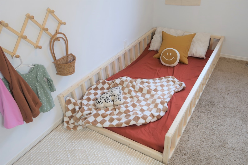 Puede incluir: Una cama infantil con estructura de madera natural y una s&aacute;bana bajera de color &oacute;xido. La cama est&aacute; adornada con almohadas, una manta a cuadros y un coj&iacute;n decorativo con una cara sonriente. Un perchero acorde&oacute;n de madera y una peque&ntilde;a cesta cuelgan de la pared.