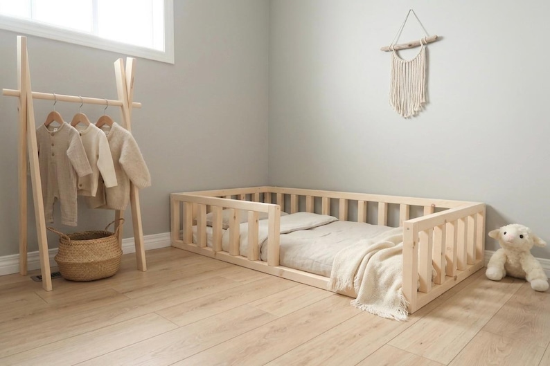 Puede incluir: Cama para ni&ntilde;os peque&ntilde;os de madera clara con laterales de listones. La habitaci&oacute;n incluye un perchero de madera con ropa de ni&ntilde;os, una cesta de mimbre y un cordero de peluche. La cama mide unos 140 cm de largo.