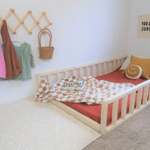 Puede incluir: Una cama infantil con marco de madera clara con una s&aacute;bana roja, una manta a cuadros y almohadas decorativas. Un estante de madera montado en la pared contiene una parte superior rosa, una chaqueta marr&oacute;n y un vestido verde. Un letrero dice "YOU ARE MY SUNSHINE".
