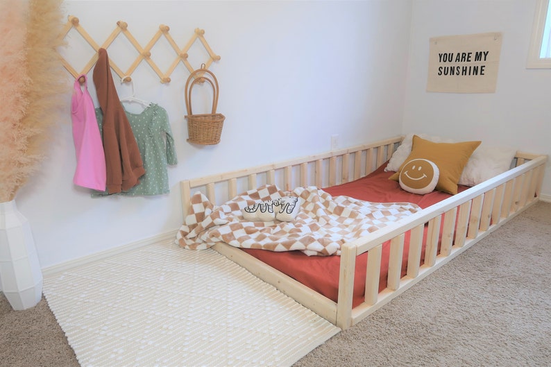 Puede incluir: Una cama infantil con marco de madera clara con una s&aacute;bana roja, una manta a cuadros y almohadas decorativas. Un estante de madera montado en la pared contiene una parte superior rosa, una chaqueta marr&oacute;n y un vestido verde. Un letrero dice "YOU ARE MY SUNSHINE".