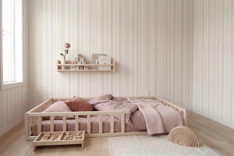 Puede incluir: Una cama con estructura de madera clara con edred&oacute;n, almohadas y manta rosas. Una peque&ntilde;a estanter&iacute;a de madera contiene libros y bloques. Un arco&iacute;ris de madera y una peque&ntilde;a bandeja de madera con juguetes est&aacute;n en el suelo. La habitaci&oacute;n tiene papel pintado a rayas.