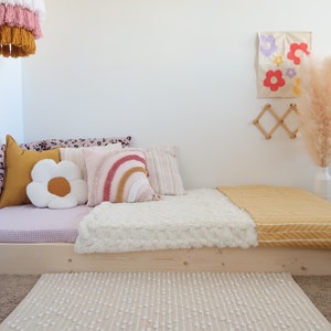 Puede incluir: Una cama baja de madera clara con varios cojines decorativos, incluyendo uno en forma de flor y otro de arco&iacute;ris. Una manta amarilla y una alfombra crema a&ntilde;aden calidez. Una l&aacute;mpara colgante y arte mural completan la decoraci&oacute;n de la habitaci&oacute;n.