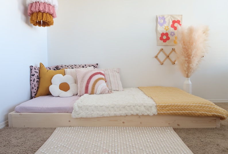 Puede incluir: Una cama baja de madera clara con varios cojines decorativos, incluyendo uno en forma de flor y otro de arco&iacute;ris. Una manta amarilla y una alfombra crema a&ntilde;aden calidez. Una l&aacute;mpara colgante y arte mural completan la decoraci&oacute;n de la habitaci&oacute;n.