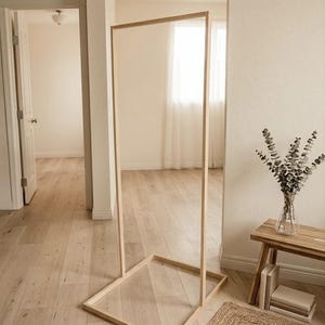 May include: A tall, rectangular wooden frame with a clear panel, standing upright on a light wood floor. The frame is positioned in a room with neutral-toned walls and a doorway visible in the background. A small rug and a wooden bench with a vase of greenery are also present.