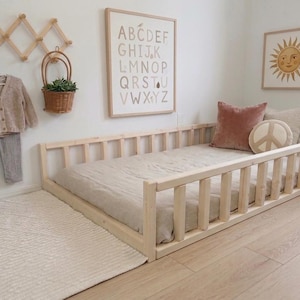Puede incluir: Una cama con marco de madera clara y colch&oacute;n beige de perfil bajo. La cama est&aacute; en la habitaci&oacute;n de un ni&ntilde;o con una alfombra, ropa colgando en la pared y arte decorativo. La habitaci&oacute;n tiene una paleta de colores neutros.