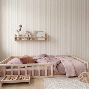Puede incluir: Una cama con estructura de madera clara con edred&oacute;n, almohadas y manta rosas. Una peque&ntilde;a bandeja de madera con juguetes se encuentra frente a la cama. Tambi&eacute;n se ve un arco&iacute;ris de madera y una estanter&iacute;a con libros y bloques.