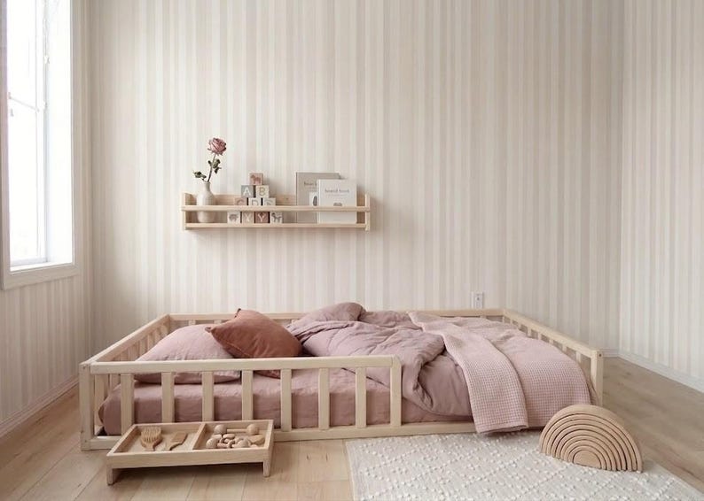 Puede incluir: Una cama con estructura de madera clara con edred&oacute;n, almohadas y manta rosas. Una peque&ntilde;a bandeja de madera con juguetes se encuentra frente a la cama. Tambi&eacute;n se ve un arco&iacute;ris de madera y una estanter&iacute;a con libros y bloques.