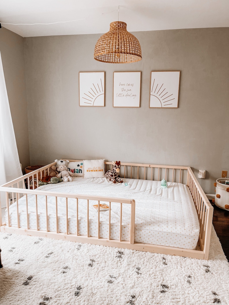 May include: A light wood framed toddler bed with a white mattress and a white and gray patterned sheet. The bed is on a white rug with a black pattern. Decorative pillows and stuffed animals are on the bed. Three framed prints hang on the wall.