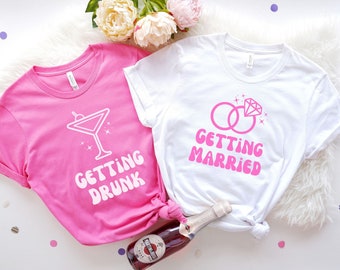 May include: Two white t-shirts with pink text and graphics. One shirt says "Getting Drunk" with a martini glass graphic. The other shirt says "Getting Married" with a graphic of two pink rings with a diamond.