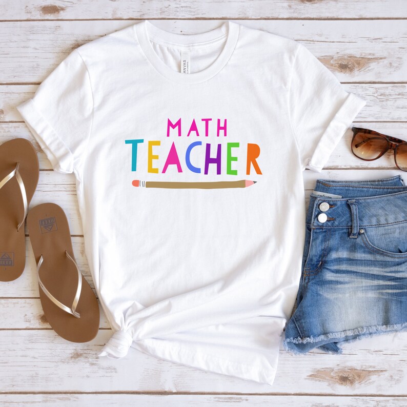 K&ouml;nnte beinhalten: Wei&szlig;es T-Shirt mit dem Text "MATH TEACHER" in bunten Buchstaben und einer braunen Bleistiftgrafik.
