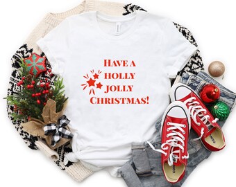 May include: White t-shirt with red text that says "Have a Holly Jolly Christmas!" with a red starburst graphic. The shirt is laid out on a white background with a sweater, jeans, and red sneakers.