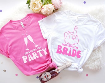 May include: Two pink and white t-shirts with the text "Here comes the party" and "Here comes the bride" in a retro font. The bride shirt features a hand with a ring on the middle finger giving the middle finger.