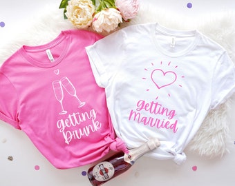 May include: Two white t-shirts with pink text. One shirt says "getting drunk" with a drawing of two champagne glasses. The other shirt says "getting married" with a pink heart.
