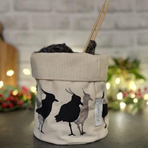 May include: A beige canvas yarn bowl with a black and white silhouette print of three birds. The bowl is filled with yarn and knitting needles.