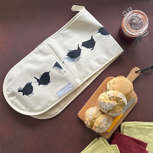 May include: A white oven mitt with a black and white design of birds. The mitt has a pocket on the side and the text "Sandra Vick" is printed on the bottom. The mitt is on a brown surface with a wooden cutting board with three round rolls of bread on it.