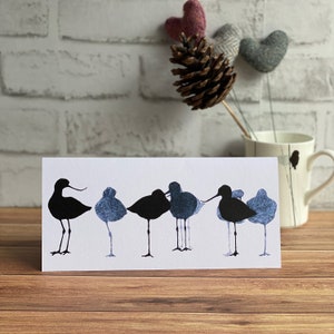 Peut inclure: Une carte blanche avec une silhouette bleue de sept oiseaux debout en rang. Les oiseaux font face au côté droit de la carte. La carte est sur une surface en bois.