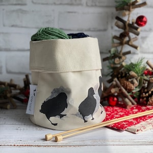 Peut inclure: Un bol à laine en toile blanche avec une silhouette noire de trois oiseaux et le mot "Écosse" imprimé sur le côté. Le bol est rempli de laine verte et bleue et deux aiguilles à tricoter en bois reposent sur un tissu rouge et blanc.