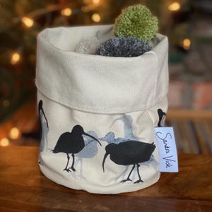 May include: A cylindrical, off-white fabric storage basket with a folded-over top. The basket features a repeating pattern of black and blue bird silhouettes. The interior holds three pom-poms in gray and green. A small tag reads "Sandra Vick".