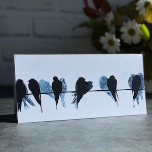 Op de afbeelding: Een witte wenskaart met een rij zwarte vogel silhouetten op een dunne draad. De vogels hebben blauwgrijze schaduweffecten. De achtergrond toont vage bloemen en een witte bakstenen muur.